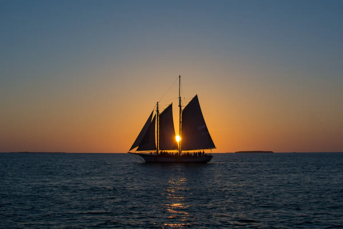 Two-masted schooner with passengers on deck, the setting sun between its sails off Key West